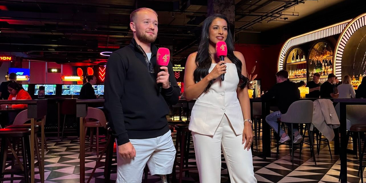 Ariana bravo stands on the left of Billy Monger, holding a pink microphone with channel 4 logo. At the F1 arcade in London, mid-way through presenting to camera.