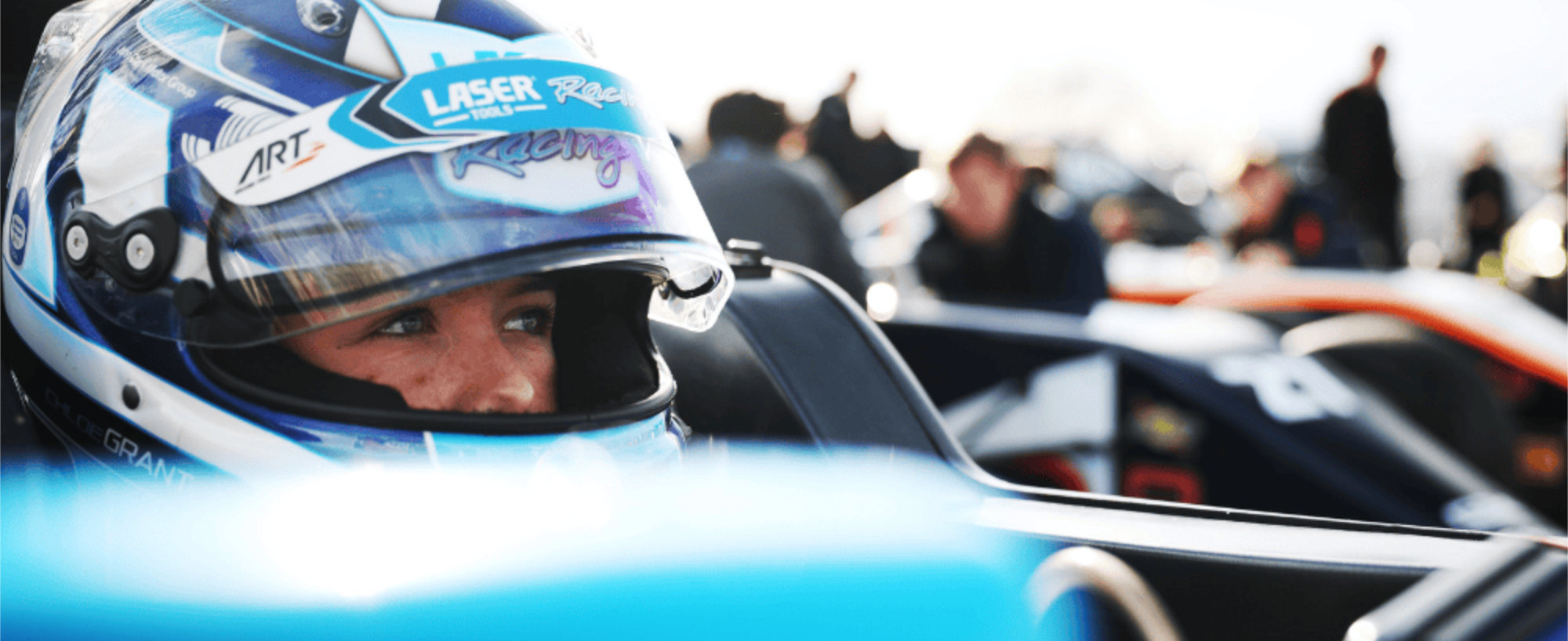 Chloe Grant in her car, close up shot of wearing helmet with visor open, looking away at camera. Other cars can be seen in the background, suggesting this is the starting grid. 