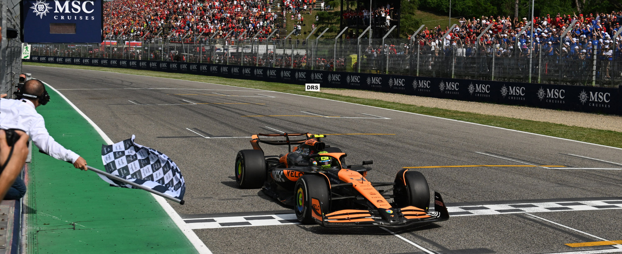 Lando Norris's McLaren Number 4 crossing the line at Imola, the chequered flag waving in the foreground. 