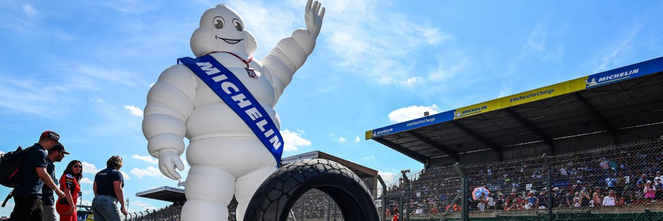 Michelin man waves to a crowd at a race track. A tyre is seen in front of him. 