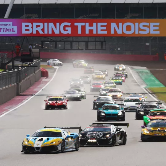 Cars on track down Hamilton Straight. Gantry saying 'Bring the Noise' is visible in the background, with a Lamborghini out in front