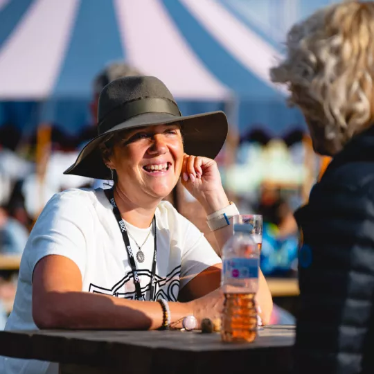 Guest wearing hat sat at a table, talking to another guest. Sunset light, with tent behind.