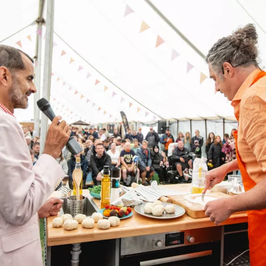 Foodie fest tent, with crowd in the background, presenter left, and chef right, preparing food.